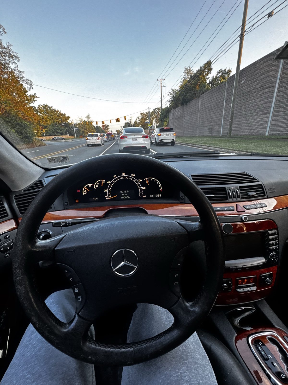 Driver's view over a Mercedes steering wheel on a tree-lined road at golden hour, the odometer reading 180,836 miles
