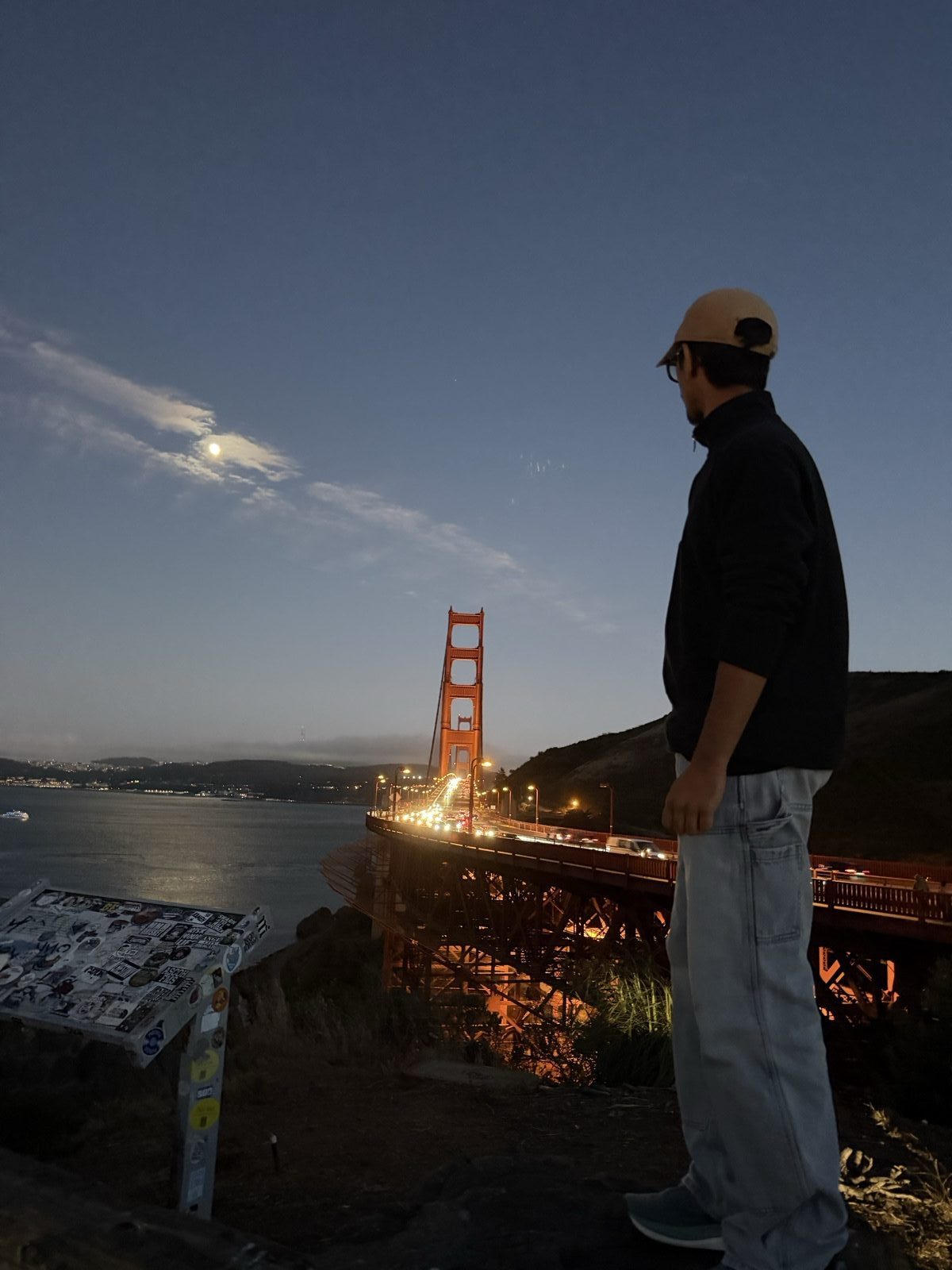 Standing on the headland above the Golden Gate Bridge at dusk, its lights on and the moon out over the bay