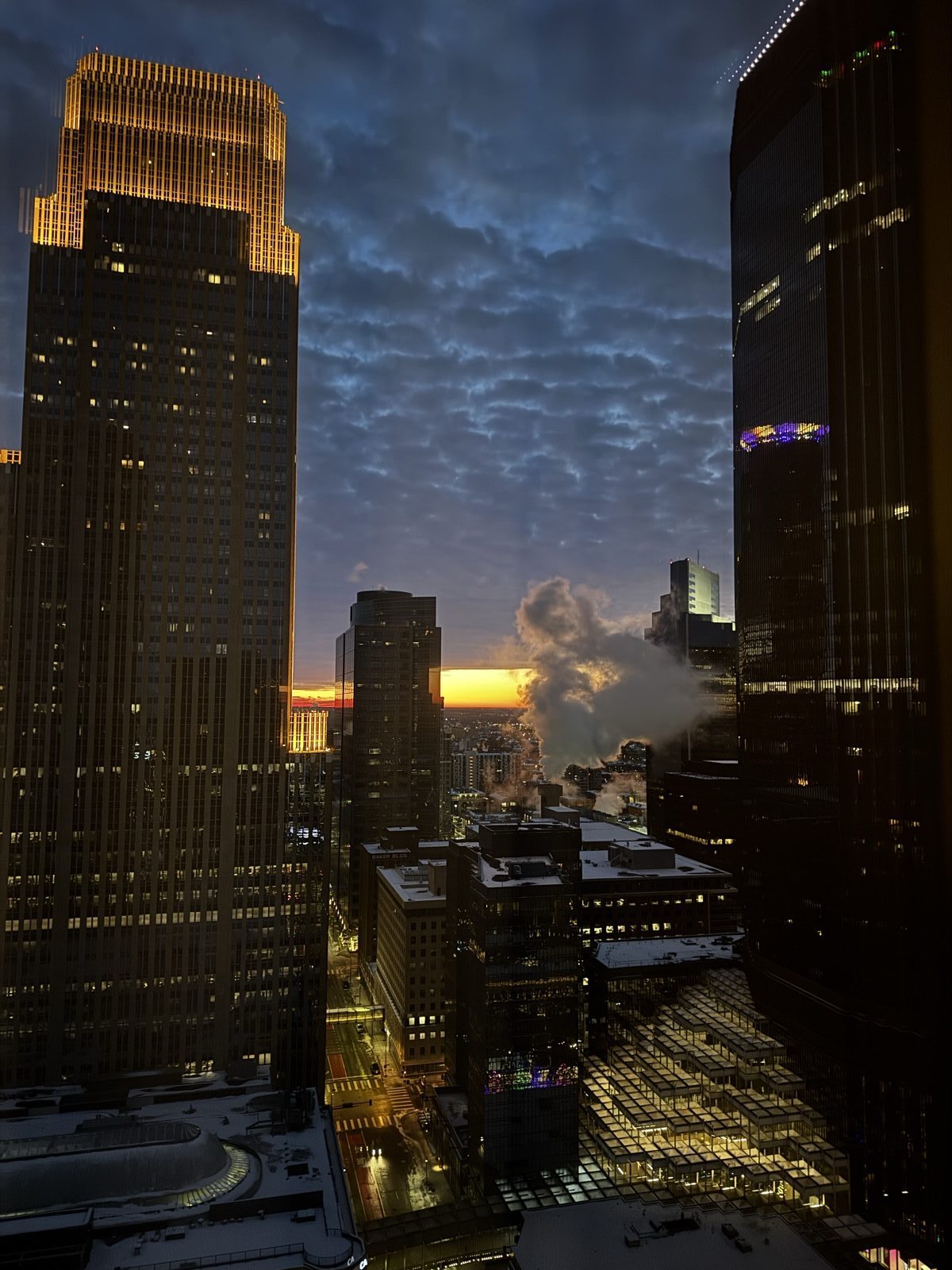 Downtown Minneapolis from above at dusk, steam rising between lit towers and the last of the sun on the horizon